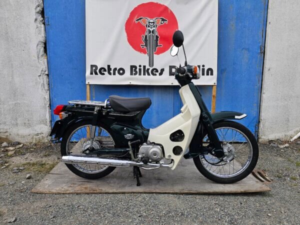 Full side view of the 2002 Honda Super Cub C50 in dark green and cream, displayed in front of the Retro Bikes Dublin banner.