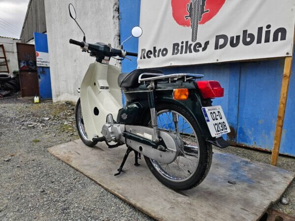 Angled side-rear view of the Honda Super Cub C50, showing the exhaust, chrome luggage rack, and rear fender.